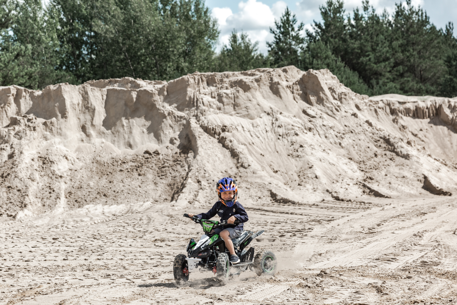 Vaikas su šalmu važiuoja smėlio karjere ant Python keturračio, tolumoje matosi kalnai