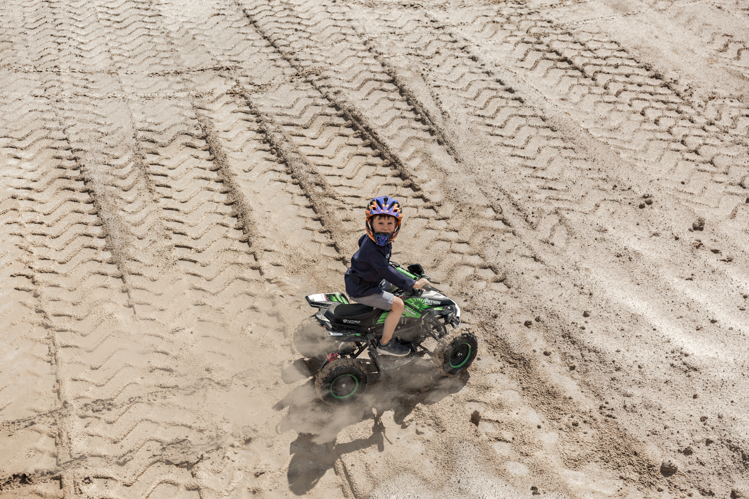 Vaikas su šalmu smėlio karjere važiuoja ant žalio keturračio, matosi ratų žymės