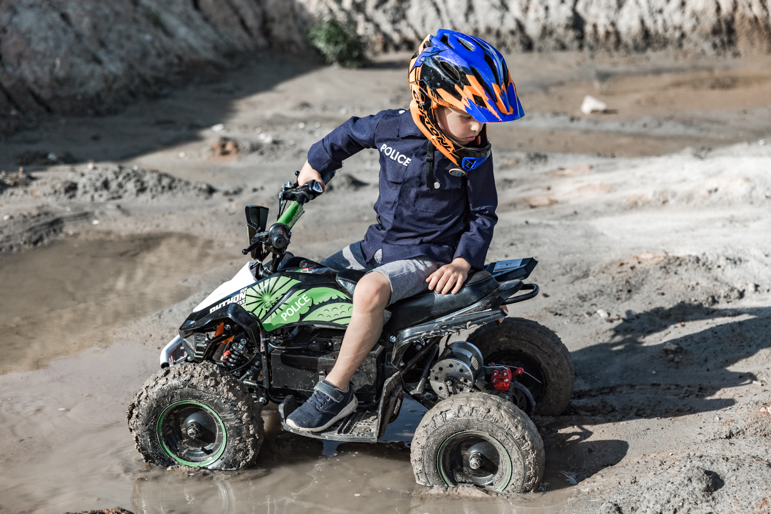 Berniukas dėvintis šalmą sėdi ant žalio keturračio, kuris įklimpęs purve