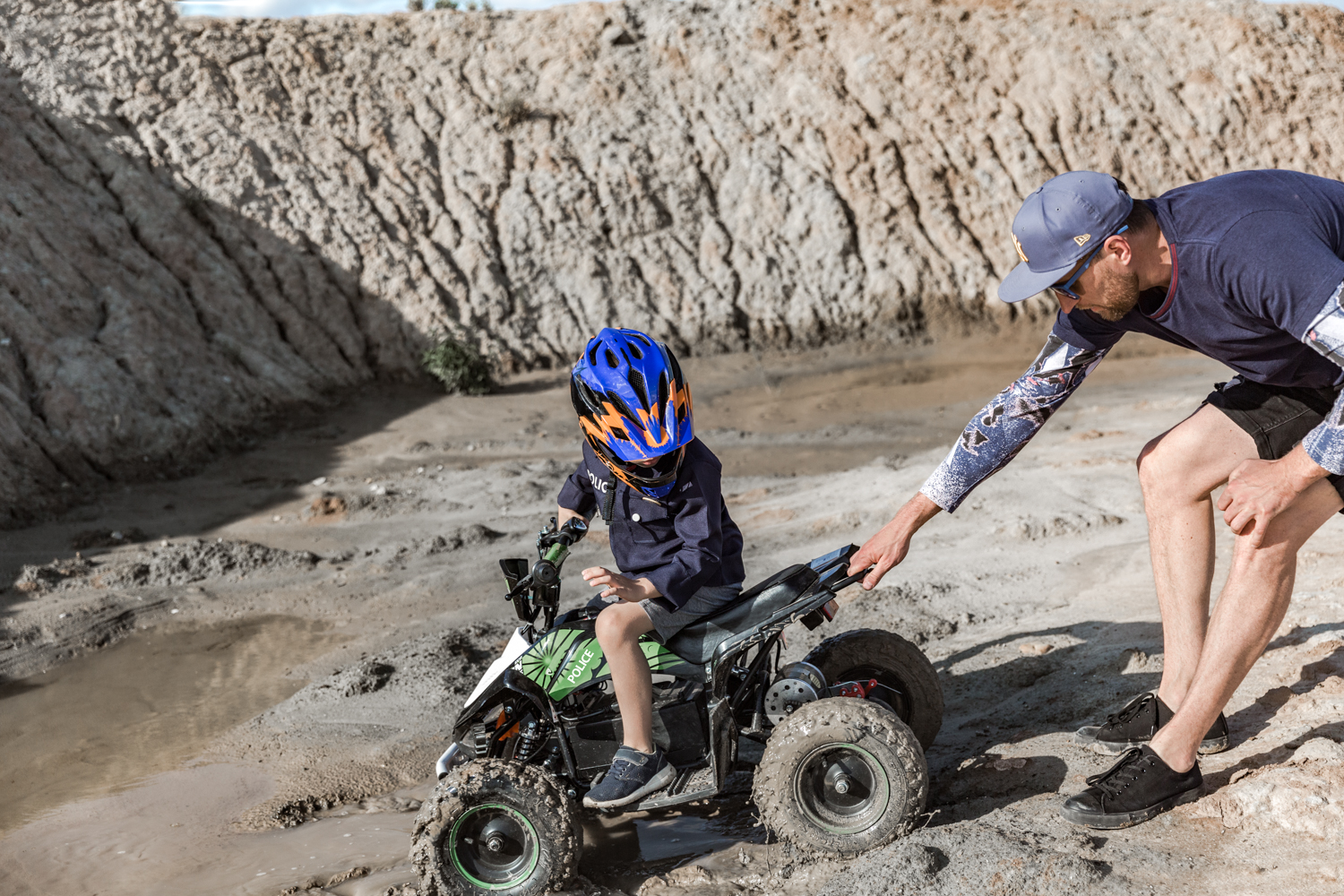 Berniukas su šalmu sėdi ant žalio keturračio, vyras tempia keturratį iš purvo