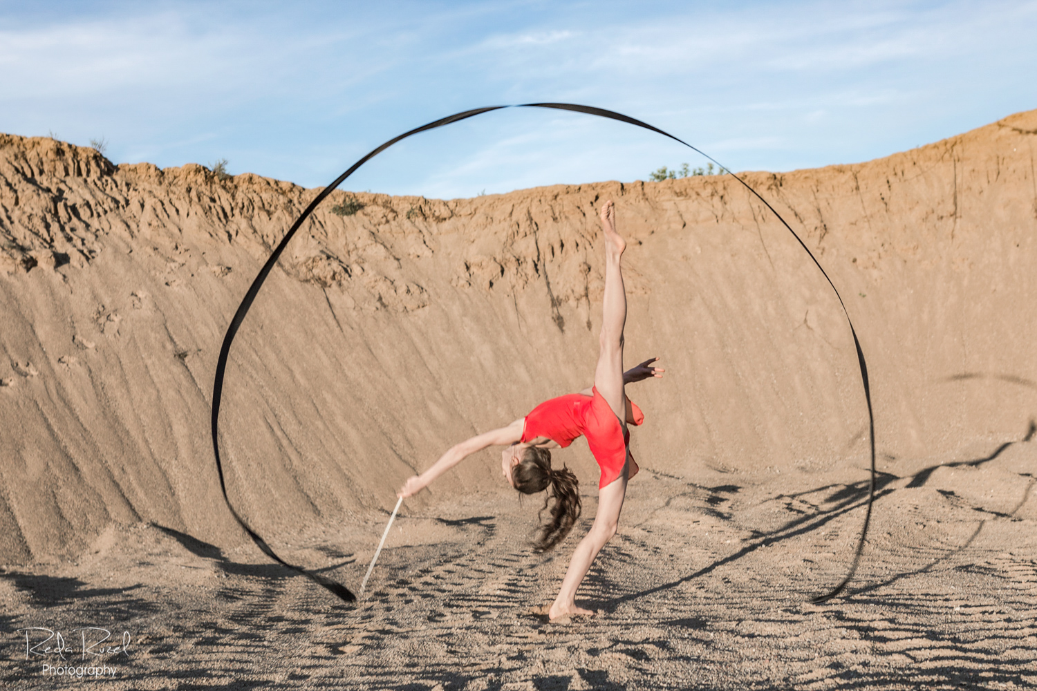 Meninės gimnastikos fotografija, mergaitė raudonu triko su juodu meninės gimnastikos kaspinu