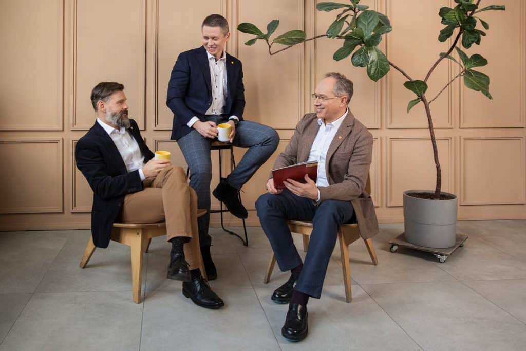 Įmonės vadovų fotosesija biure: trys vyrai kostiumais prie šviesios sienos su augalu