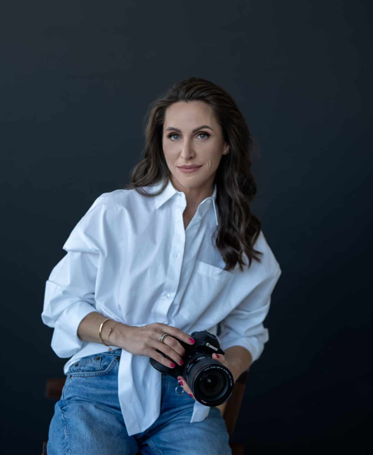 Fotografė Reda Ruzel su fotoaparatu studijoje, baltais marškiniais ir džinsais