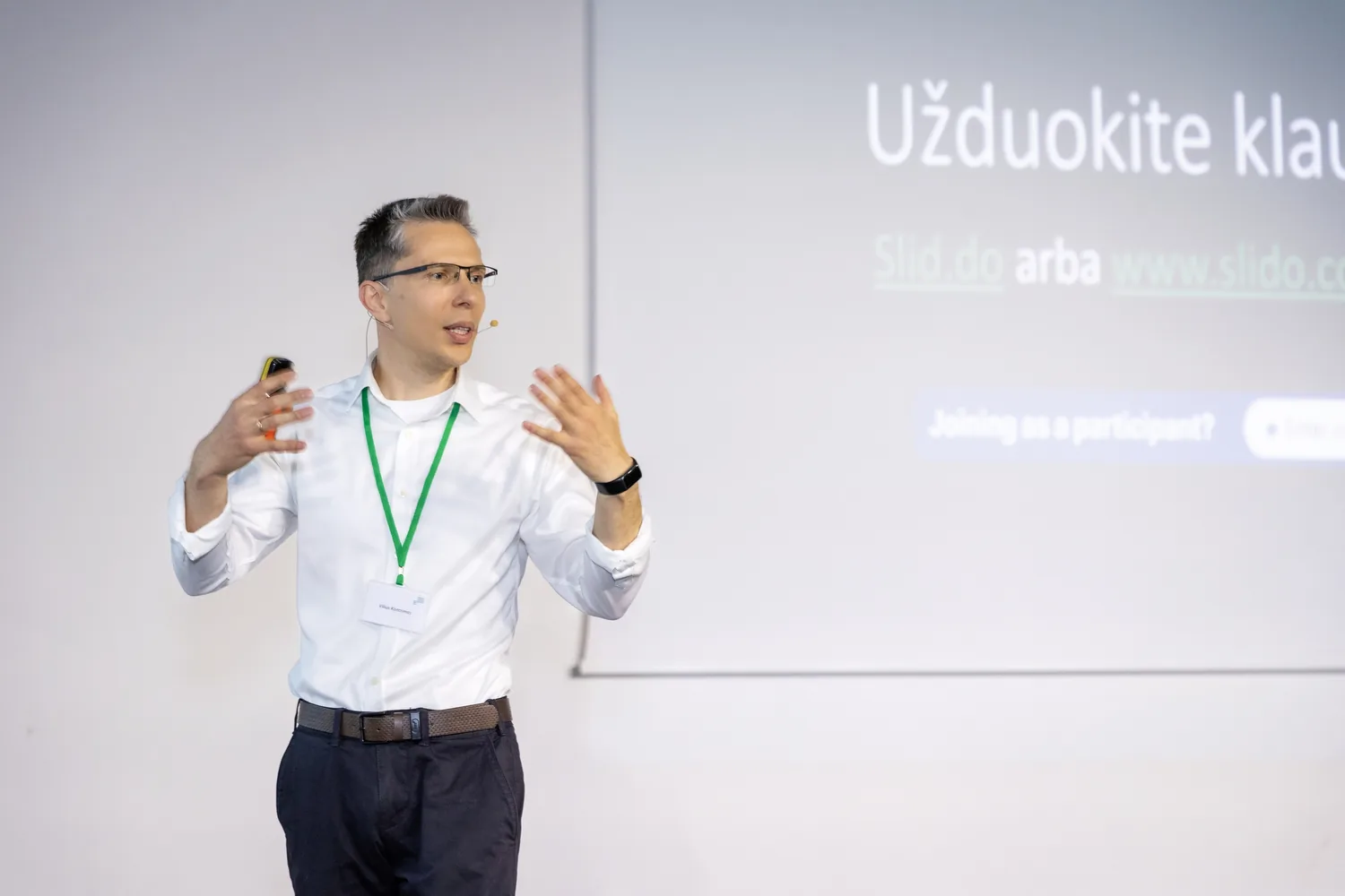 Pranešėjas su mikrofonu rankiniu gestu pasakoja konferencijos dalyviams prie pristatymo ekrano