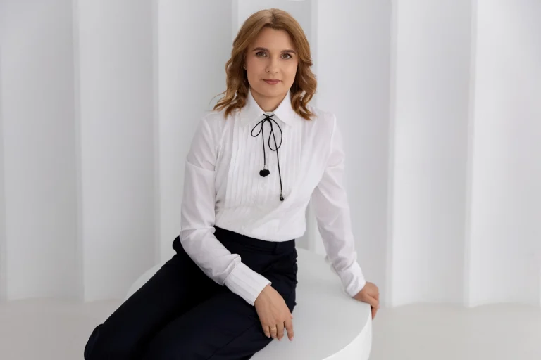 Specialistės portretas: moteris baltais marškiniais su juoda juostele baltoje studijoje